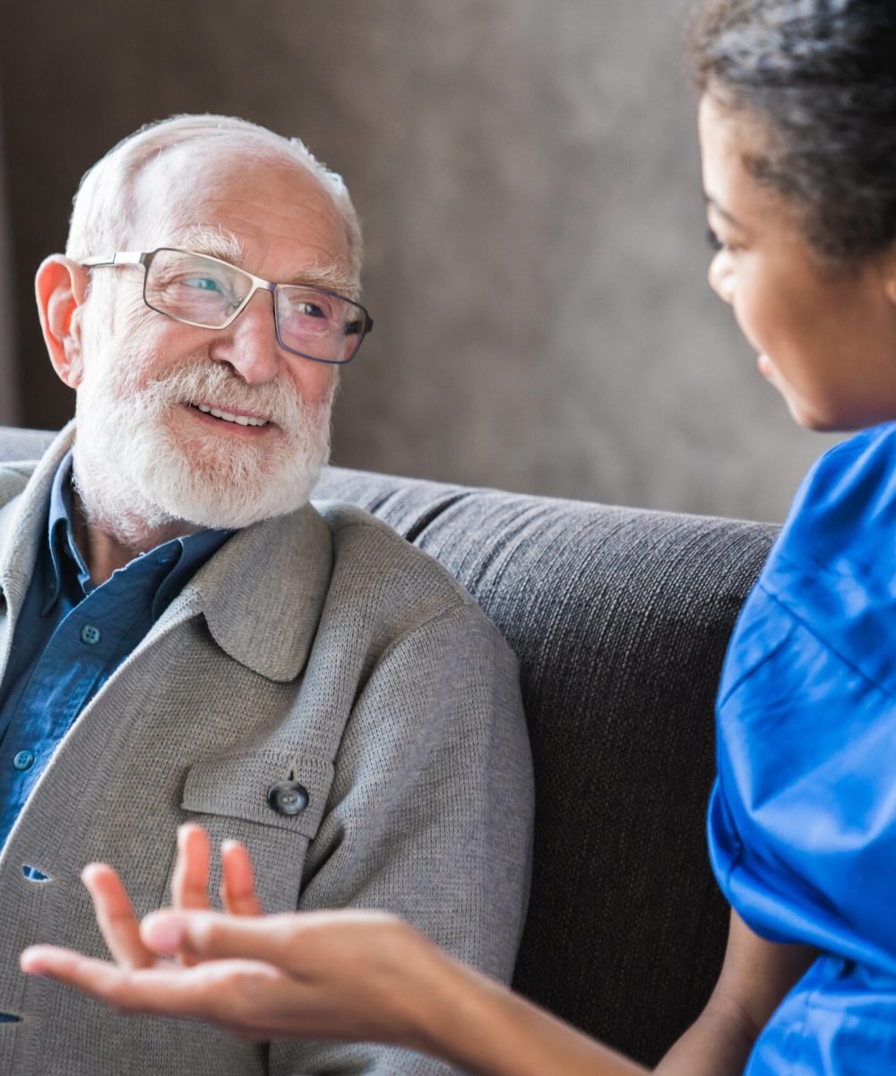 Female african professional doctor consulting senior patient grandfather during medical care visit. Young woman physician and old man talking providing medical assistance sitting on sofa. Elderly people home care concept